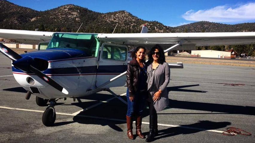 Two women smiling in front of a light aircraft on a runway, with Californian-looking landscape behind them