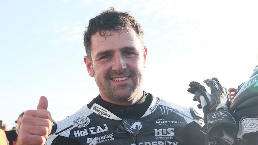 Michael Dunlop has short dark hair, he has his thumbs up and is smiling at the camera. He is wearing his black racing leathers with sponsors on it in white.