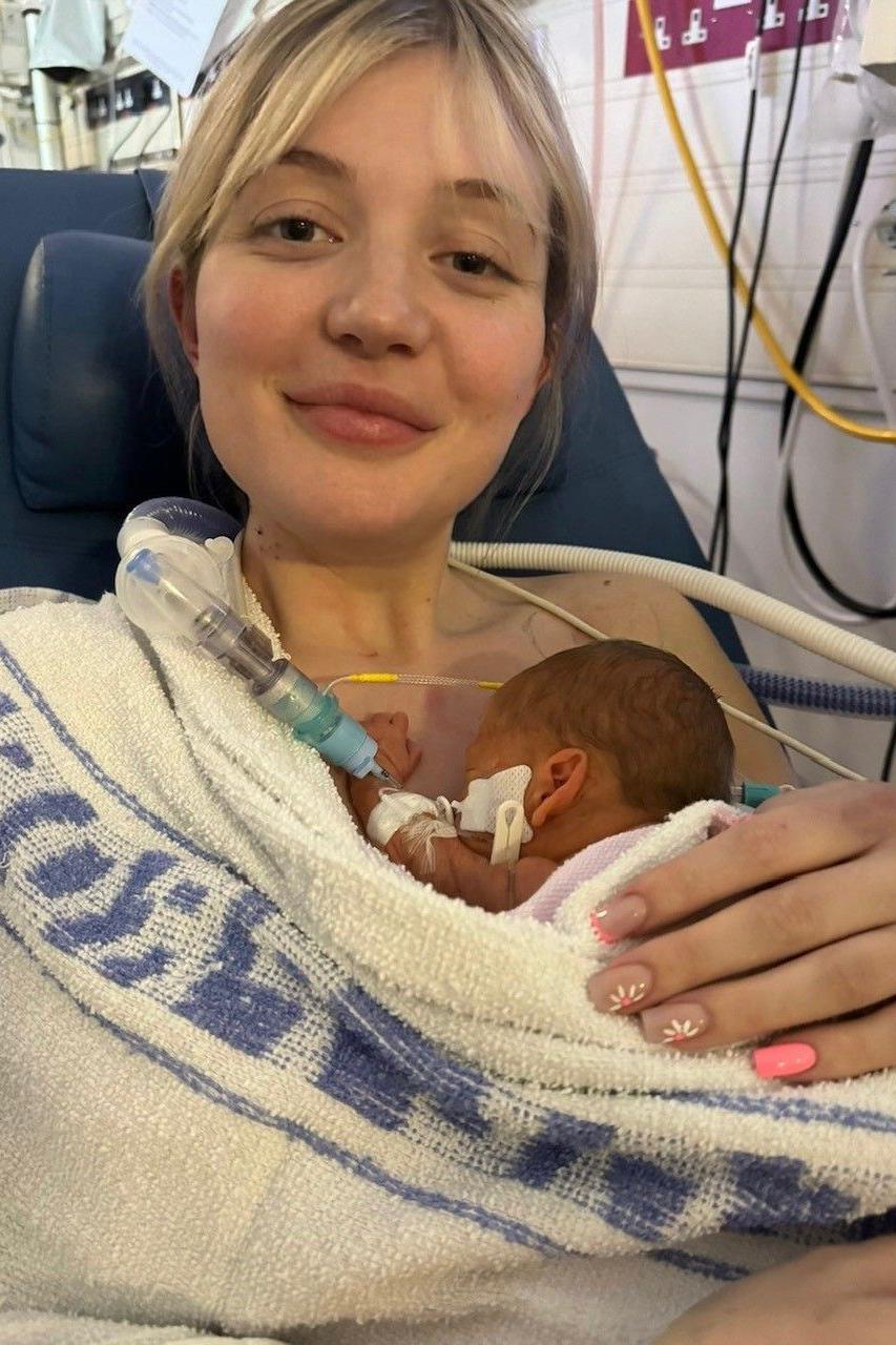 A young mother holds her newborn baby, wrapped in a hospital blanket.