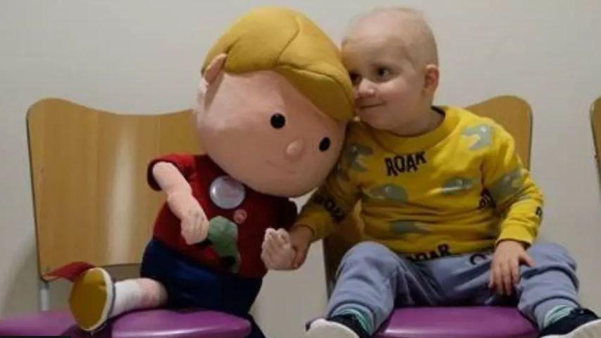 A three-year-old Ralph, who wears a yellow top with dinosaurs on it, holds hands with a doll version of himself in a hospital waiting room