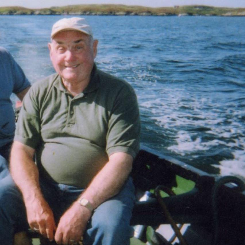 Calum is sitting in a boat and is smiling. He is wearing a green T-shirt and a beige cap. The sea is blue behind him.