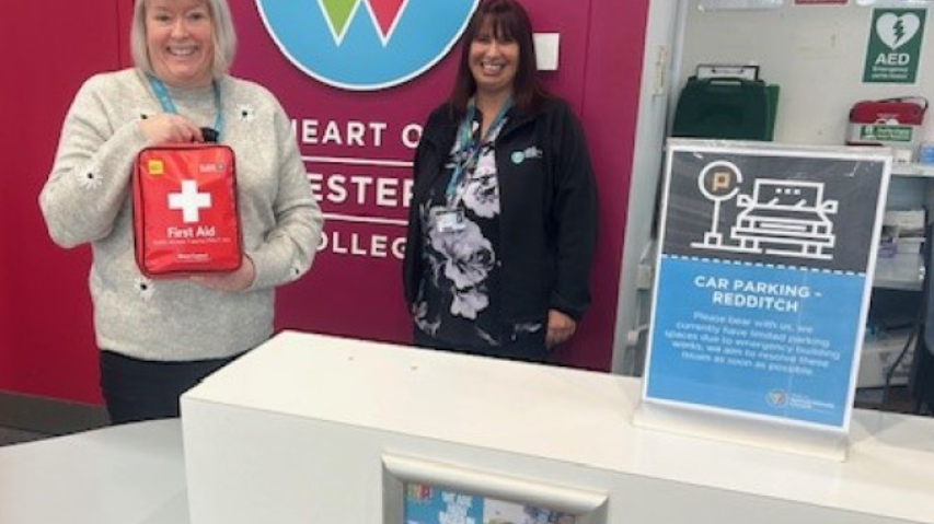 Two women are standing behind a desk, one is holding a large red bag in both hands, with a large white cross and the words first aid on the front of the bag.