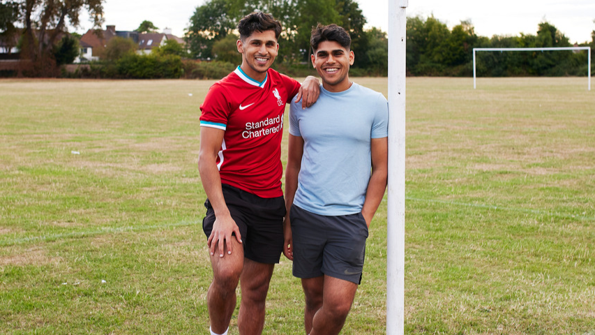 Two men of South Asian heritage standing on an empty football pitch. Both of them are smiling at the camera. 