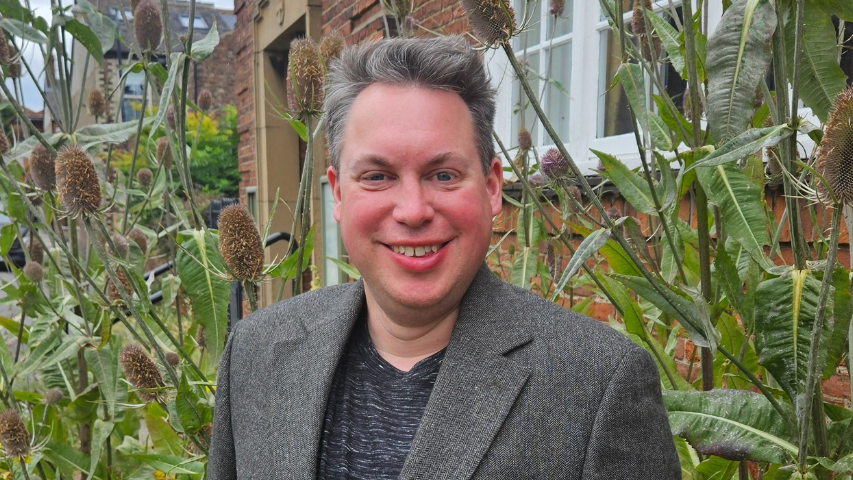 A man with short grey and white hair wearing a grey suit and T shirt smiles at the camera. Behind him are tall thistle plants.