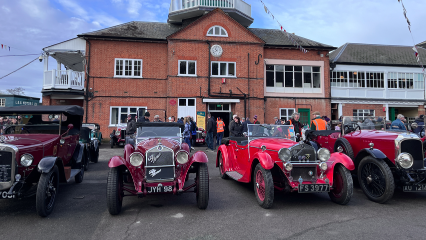 Four red vintage motors are parked in front of Brooklands Museum - a red brick building with a white viewing platform on the side.