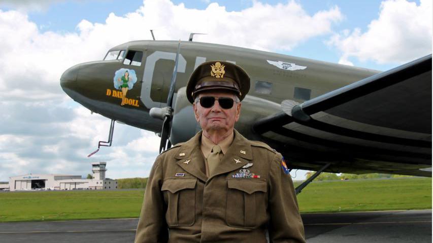 Lieutenant Colonel David Hamilton in military uniform standing in front of a military plane on a runway. He is wearing dark sunglasses