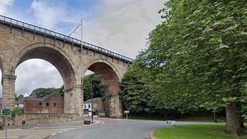 The beige stone viaduct rises above Durham's A690 roundabout with a large tree in the middle. A sign on the left points towards Crook. There a few houses and a pub further up the road. 