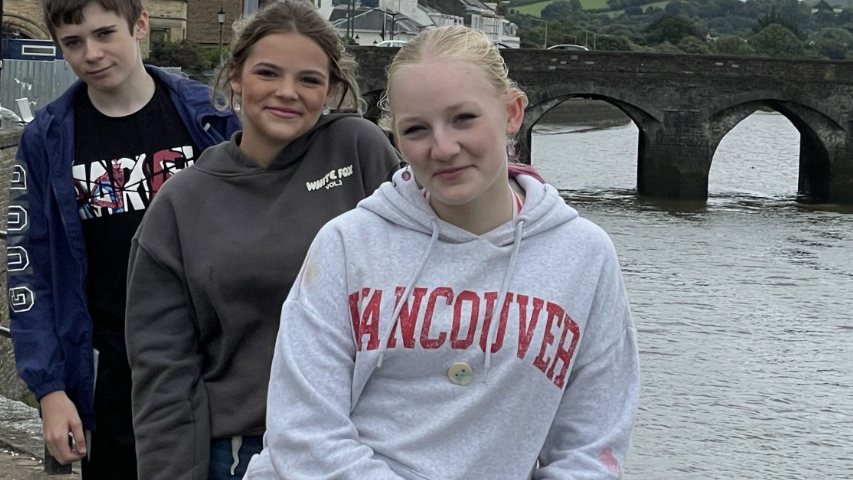 Three young people wearing blue, brown and grey hoodies with writing on then in front of a river and a bridge.