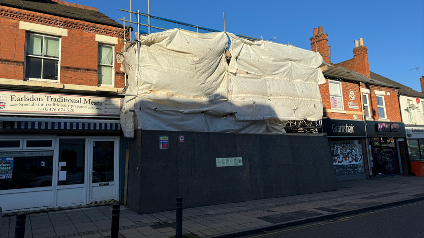 A row of shops, with white scaffolding covering two buildings in the middle. There is a gap between the buildings showing demolition work has taken place on the two in the middle.