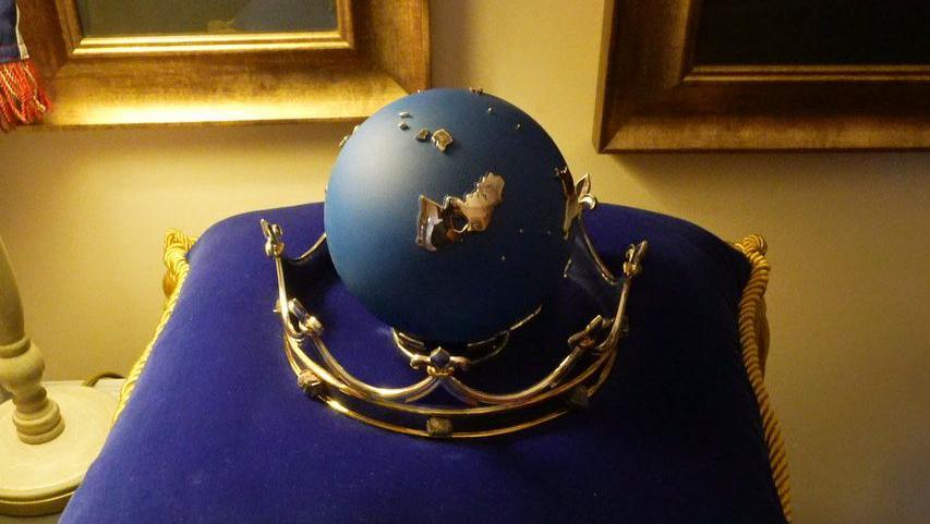 The navy-coloured globe is set in a golden metal crown. The item sits on a purple velvet cushion. The room is lit by a lamp and pictures in gold frames are on the wall behind it.