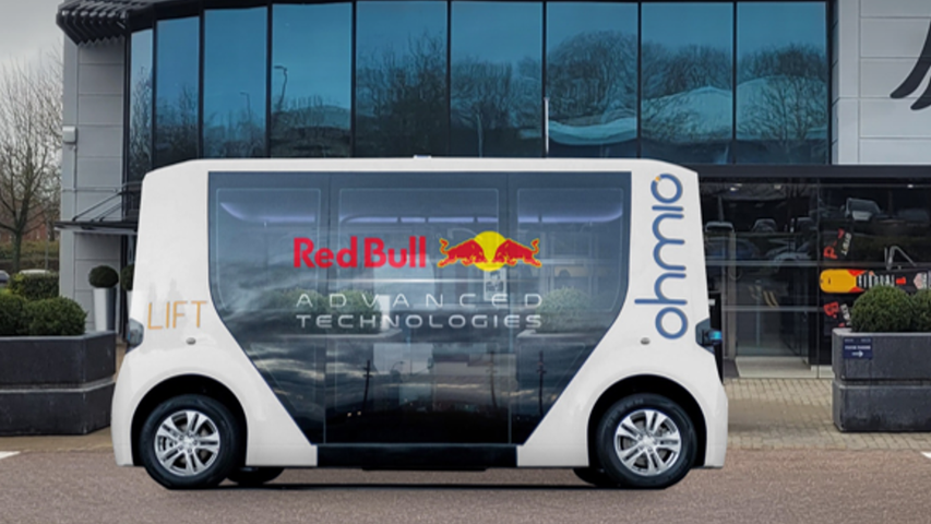 A driverless bus parked on a forecourt. It is a shuttle and is white. It has both Red Bull and Ohmio branding on it