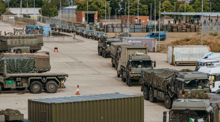 A convoy of army vehicles entering a space in single file, with various shipment containers in the area