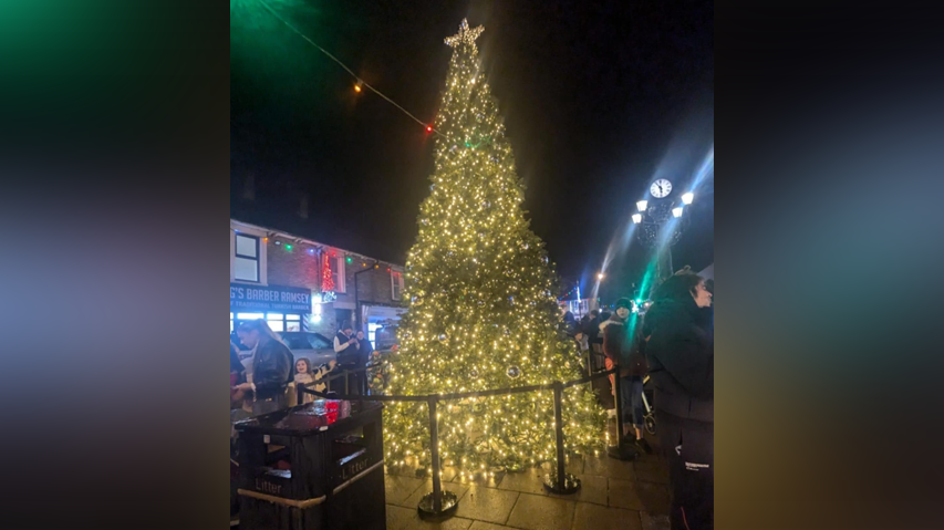 A night-time scene of a very sparkly Christmas tree with a star on top, lit up. A lightweight webbed cordon is around the tree, with shops lit up in the background