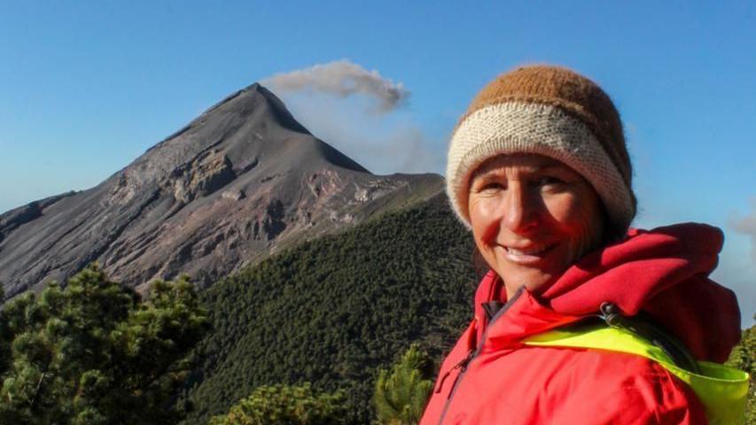 A photograph of a woman smiling wearing a red jacket and a hat. There is a volcano mountain behind her and blue skies. 