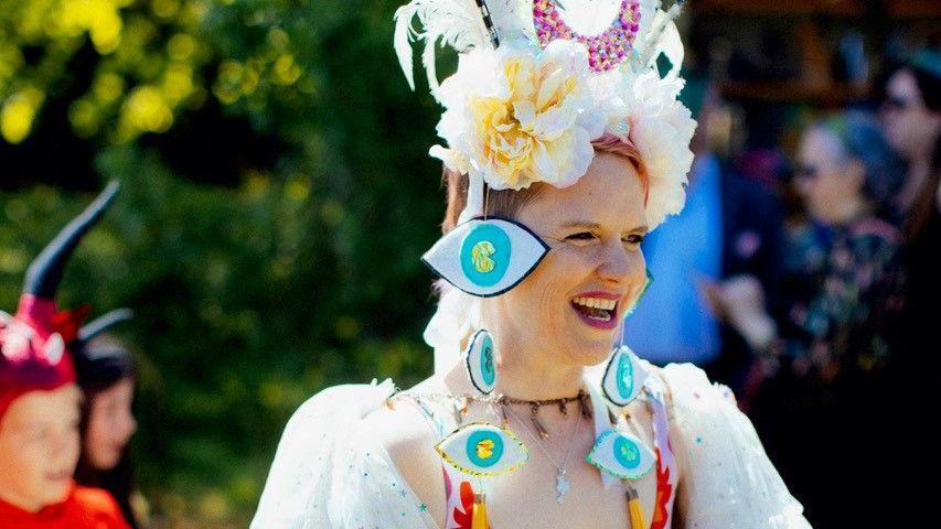 A white woman with flowers in her hair, eye shaped hair pieces, and a white dress with jewellery smiles.