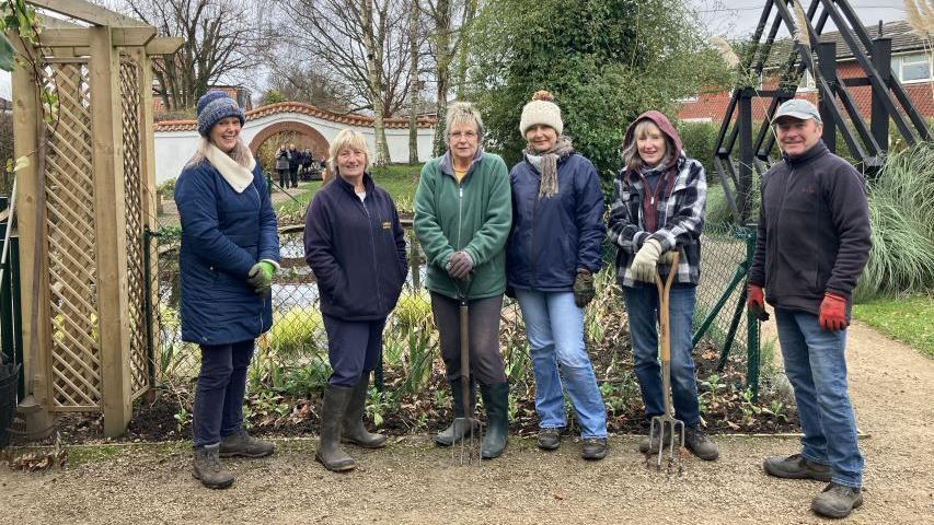 A group of six volunteers - five women and a man - standing in front of a pond. They are wearing boots and wellies, jeans and gardening gloves and two of the women are holding rakes. 