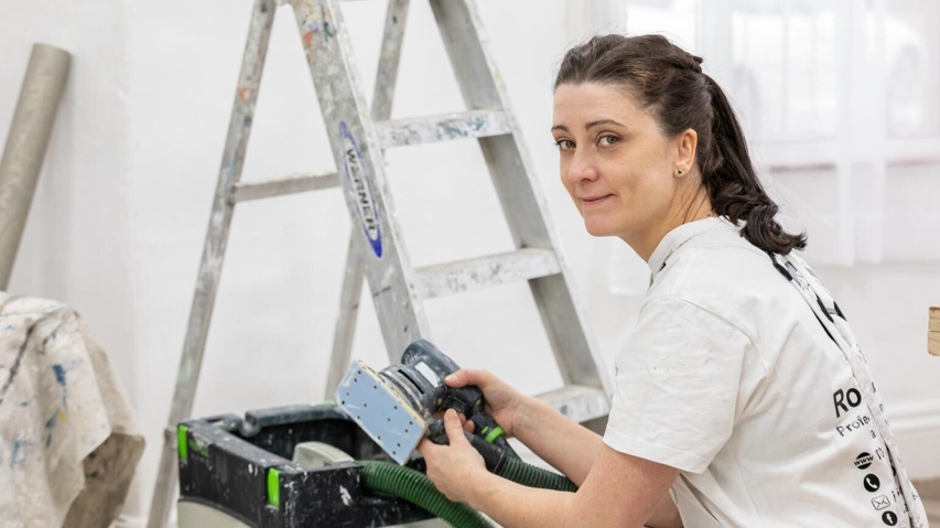 Rosie Russell in work clothes, crouching down by a ladder and holding a sander