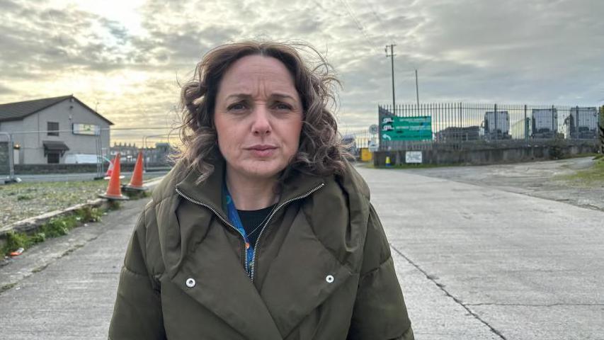 Llinos Medi looks concerned as she stands on a side road by Holyhead port, with some HGVs parked up in the background. She is wearing a camo green coat and a black top. She has curly blonde shoulder length hair.