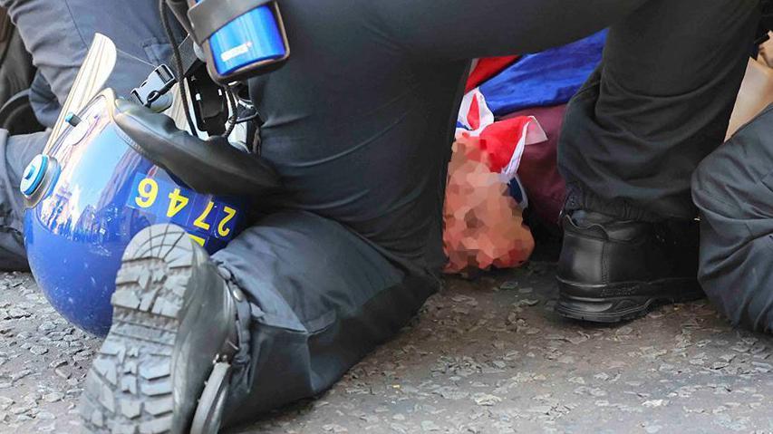 A man with his face blurred and draped in a Union Jack is pinned to the ground by two police officers