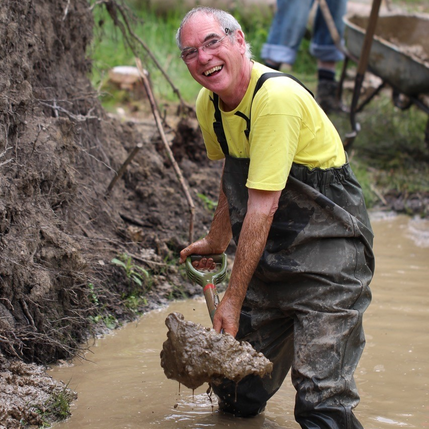 A man in a yellow T-shirt and waders is standing up to his knees in muddy water while shoveling mud onto a bank. He is wearing glasses and grinning while he works.
