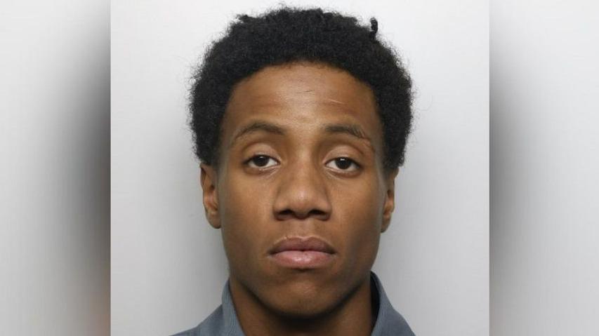 Romaine James' mugshot, where he is looking at the camera with a serious expression in front of a grey background. He has black hair.