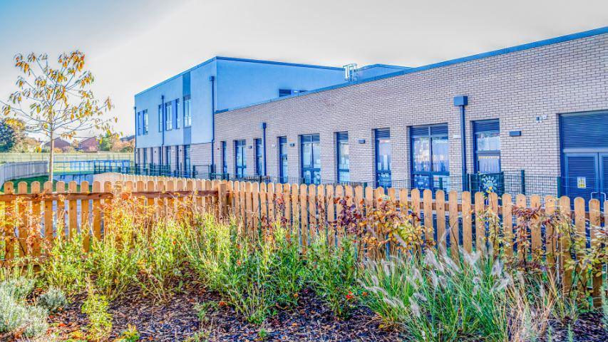 A school building with a wooden picket fence and plants and a tree in front. 