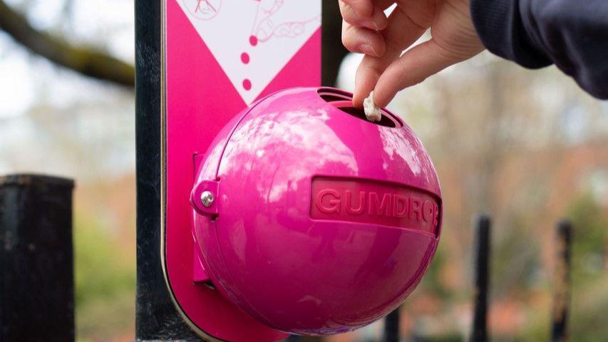 A pink, ball shaped bin on a black railing, with a circular hole on the top. Someone can be seen emptying chewing gum into it 