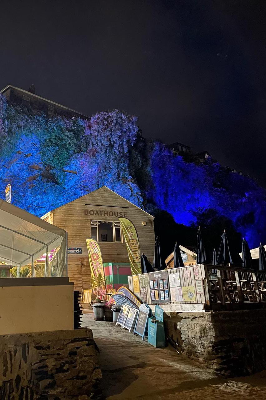 The ivy-covered wall of Newquay Harbour is lit up with blue and purple lights. In the foreground is a building with the letters BOATHOUSE on it, two flags advertising Cornish Rattler and a row of wooden chalk board signs and signs above promoting businesses in the harbour. On the left is a marquee and on the right is a raised area covered in wooden chairs.