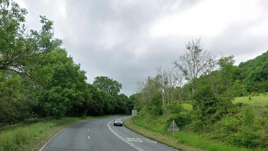A three lane road swings through green countryside. A car drives towards us on one side of the road with the word "Slow" written on the road ahead of it. The other side is made of a lane with an overtaking lane in the middle with dotted white lines between them.