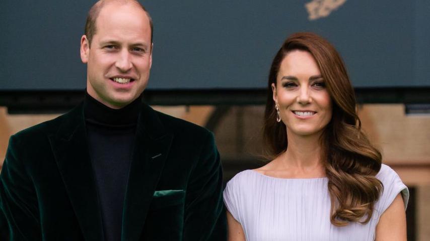 Prince William and the Duchess of Cambridge standing on the green carpet at the Earthshot awards in 2021. William is wearing a dark green velvet jacket and a navy blue roll-neck sweater. Kate has a simple white dress on. Both of them are smiling at the camera.