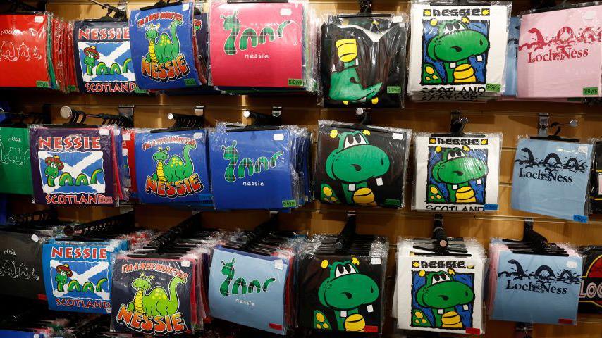 A wall of t-shirts in plastic hanging on rails. The t-shirts have different images of the Loch Ness Monster and are different colours. Most of the images show Nessie with three humps.