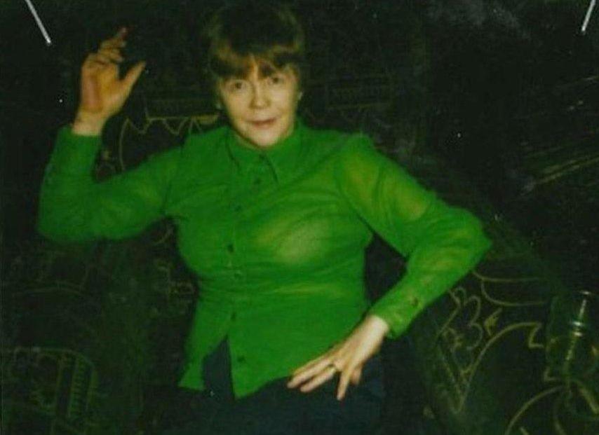 A young Mary McLaughlin poses while she dances, one hand on her hip, one in the air. She has short brown hair and is wearing a bright green chiffon shirt. She looks to be in a bar, with a dark background.