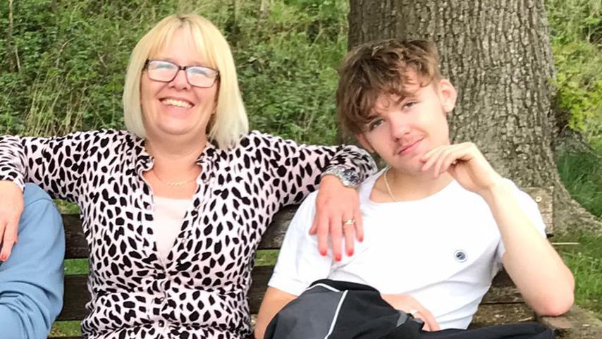 Samantha Nunn sits on a bench with her son Joe. They are sitting in front of a tree. Ms Nunn is wearing glasses, a leopard print top and blue jeans. Joe is wearing a white T-shirt. Ms Nunn is smiling at the camera with her arm on Joe's shoulder. There is another person partially in view sitting on the other side of Ms Nunn who is wearing a blue jumper. 