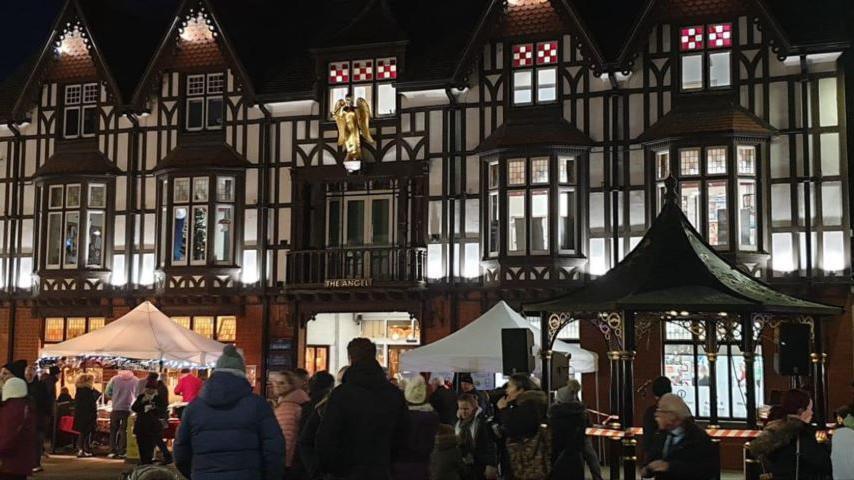 A photo of a lively night market in front of a black-and-white Tudor-style building with stained-glass windows and a golden angel statue. People wearing clothing appropriate for winter can be seen browsing stalls under white tents.