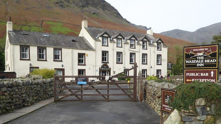 Exterior of the Wasdale Head Inn. It is painted cream and is surrounded by hills. The building is shaped like two large houses. There is a sign showing the pub's name outside. Its gate is closed and it is surrounded by a wall made of stones.
