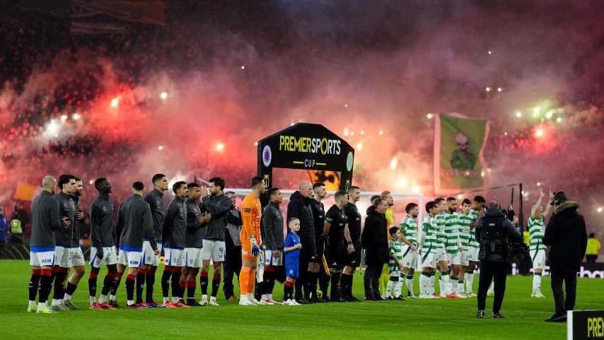 A group of Celtic and Rangers players stands on the pitch, united and focused, moments before the kick off. In the background red flare and smokes bombs that have been let off in the crowds are visible.