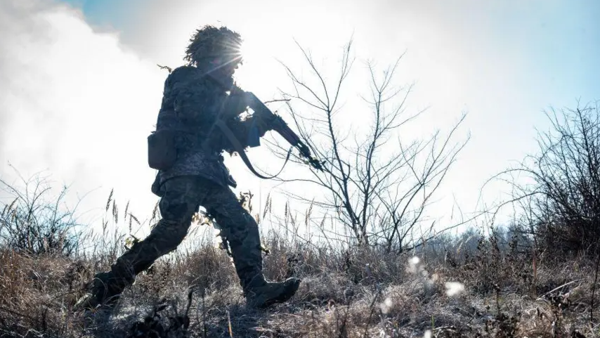 A soldier in uniform and helmet advancing across a battlefield with his gun at the ready on his shoulder. The landscape looks wintry. There appears to be frost on the ground and trees and shrubs have no leaves. The soldier is partly silhouetted by the sun, which is shining directly at the camera.