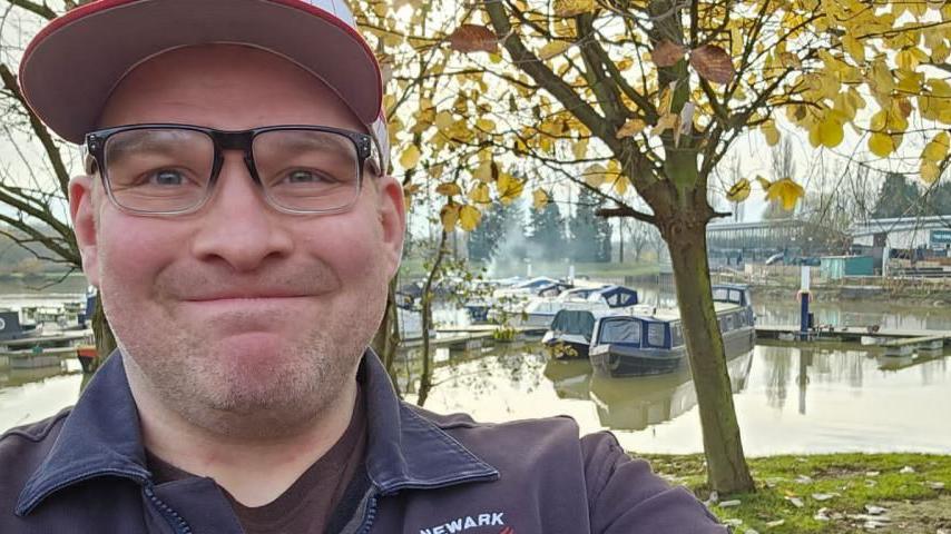 A man in a navy coat, glasses and red cap, takes a selfie by a river with narrow boats behind him. 