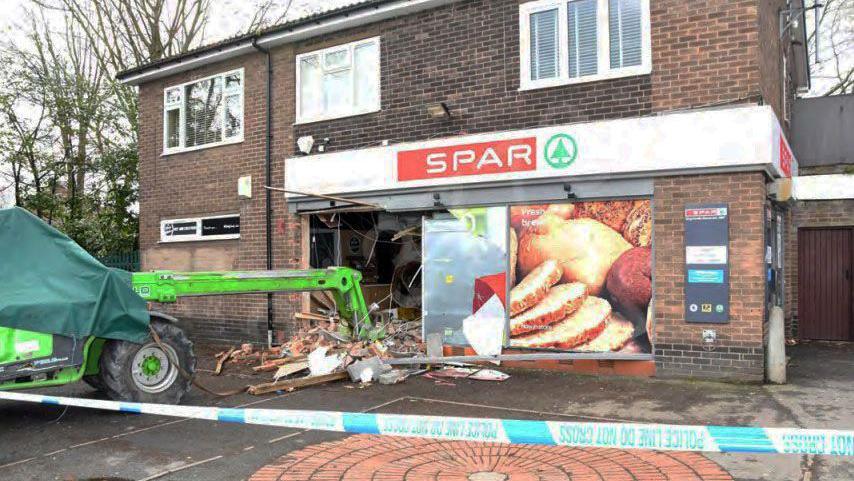 A green telehandler - a large hydraulic lifting machine - lies in the rubble of a damaged shopfront