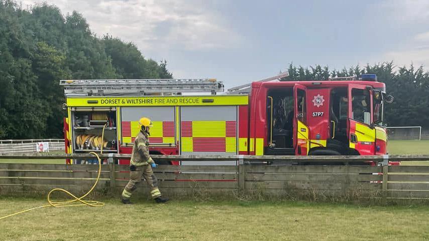 A Dorset and Wiltshire Fire and Rescue engine parked alongside a wooden fence on the football pitch. There is a firefighter wearing their kit stranding beside the fire engine, while a yellow hose comes out the back and stretches across the grass 