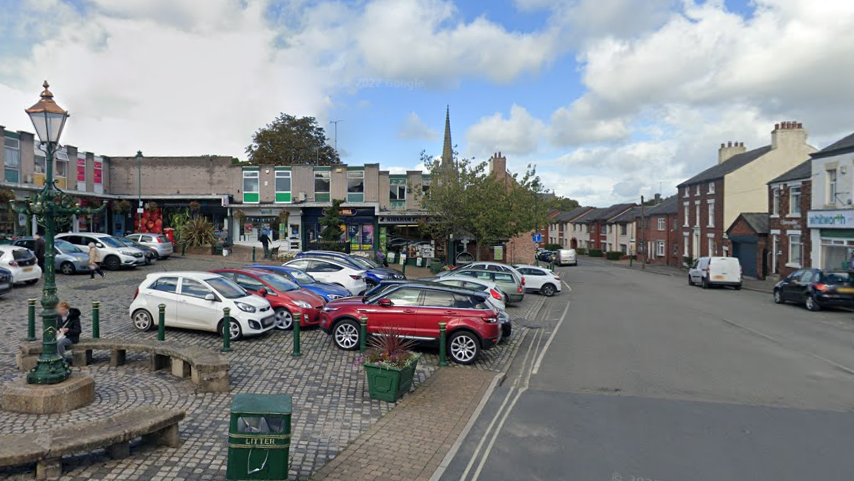 Market Square in Kirkham. The parking area is cobbled with room for about 20 cars. There are rows of shops to the left of the square, and a road and some houses and more shops to the right. There are curved benches in front of the car park for shoppers to sit on.