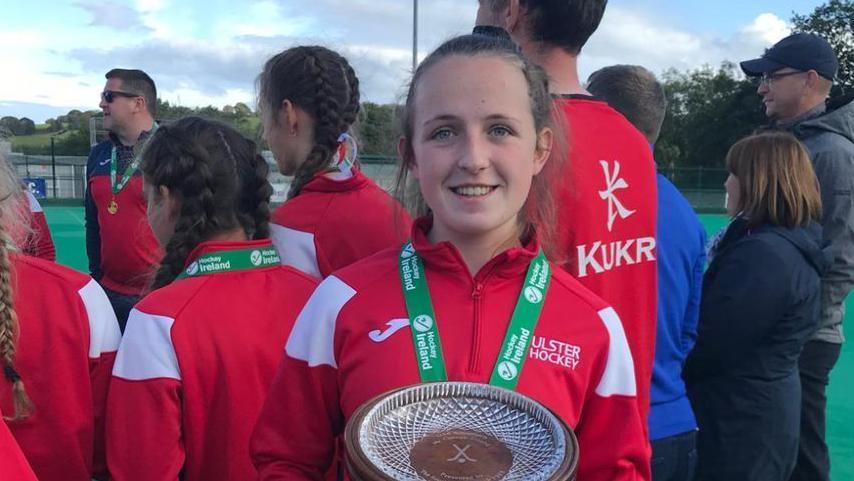 Amy with her hair pulled back wearing a red and white shell jacket which says ULSTER HOCKEY on it in white letters on the left breast. She is hold a circular crystal trophy and has a medal around her neck on a green ribbon.