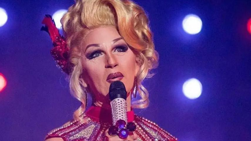 Head and shoulders shot of Zoe in full drag in a blonde wig, hair piled high and holding a microphone against a blue background with show spotlights