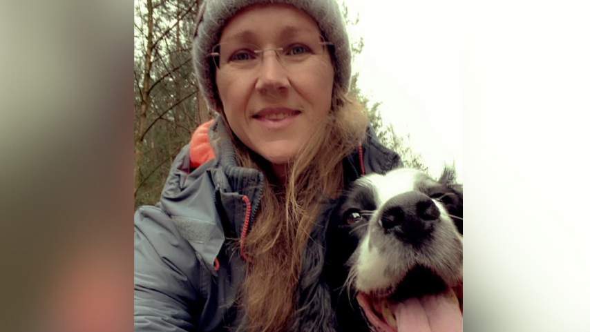 Mel Harrison wearing a hat and coat while standing with her dog in a wooded area. Her dog is a white and black spaniel and has its tongue hanging out.