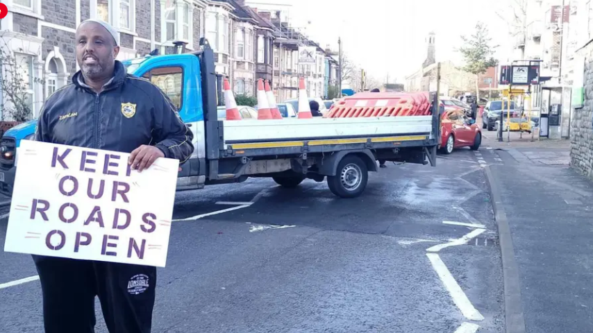 A man standing in the street with a placard saying "Keep our roads open". A contractor's lorry is behind him with road cones and barriers in it, and terraced houses stretching into the background