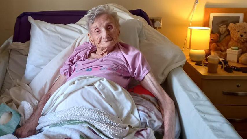 An elderly woman with short grey hair lying in a bed smiling. She's wearing a pink top. The bedside table next to her has a lamp, a teddy bear, a picture of a man and a cup.