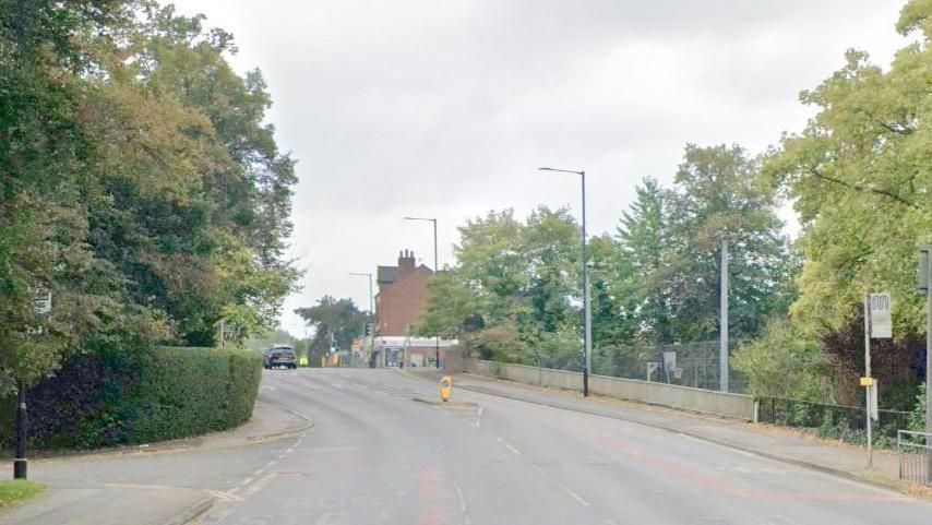 Trees line the road on Bramhall Lane with some houses seen in the background.