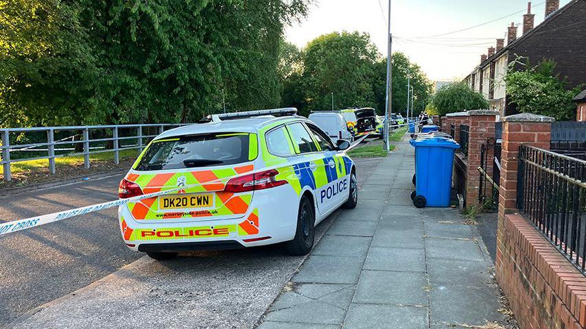 Merseyside Police vehicles inside a cordon with a row of houses on the right of Tobruck Road in Huyton and trees to the left lining the road on a sunny evening.