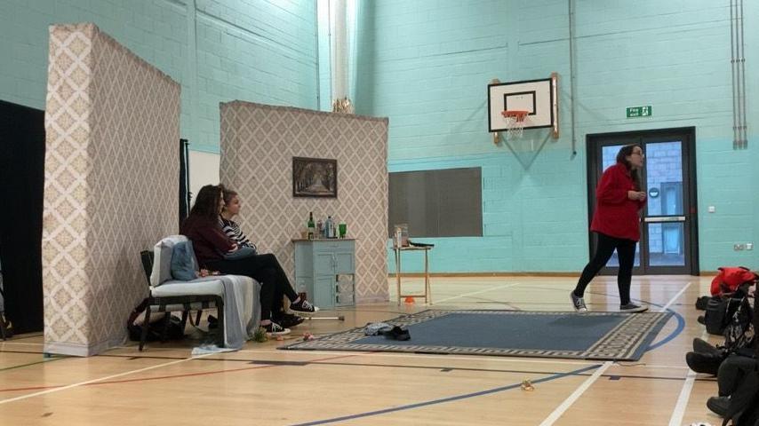 Two women sitting down on a chair and another woman speaking to students at the Last Orders performance at St Sampson's High School