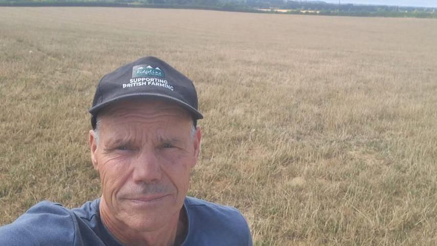 Duncan Palmer, a middle-aged man wearing a baseball cap is stood in a field of yellow grass.